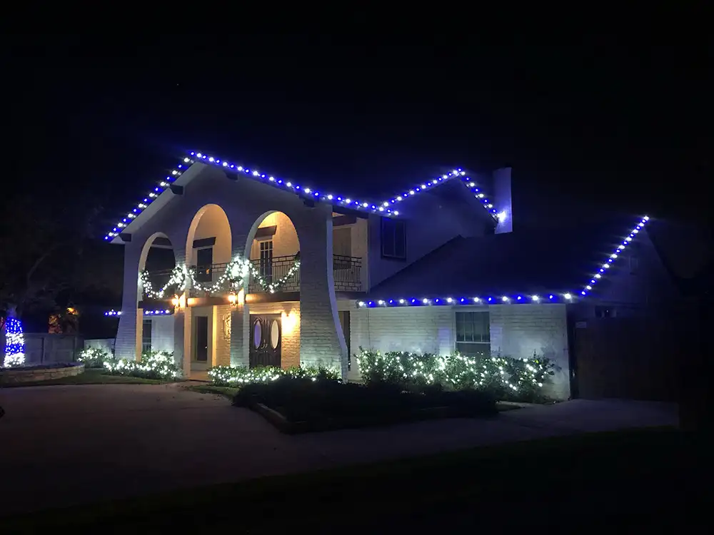 Blue and white Christmas lights outlining rooflines, garland, and shrubs on a two-story home at night