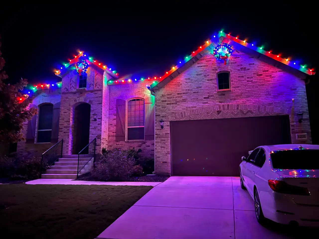 Multicolored Christmas lights outlining rooflines and wreaths on a brick home at night