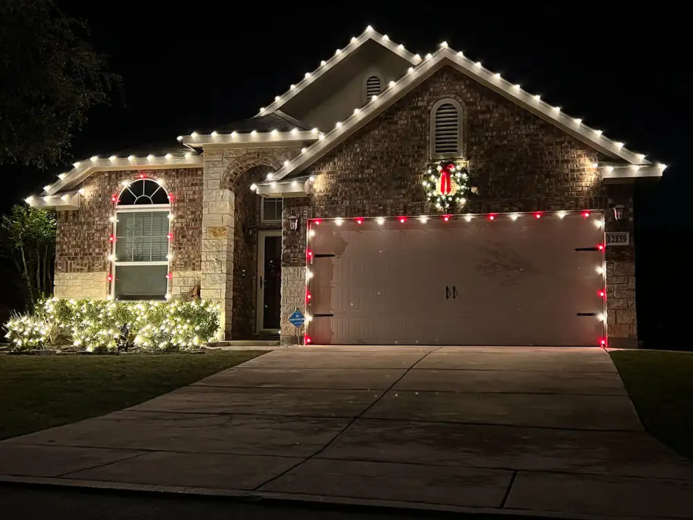 Brick home with white roofline Christmas lights, red garage outline, and lit wreath at night