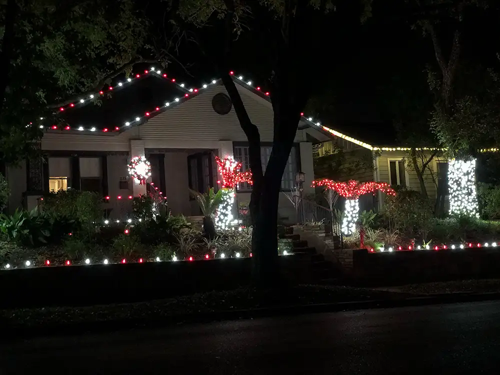 Bungalow with red and white roofline Christmas lights, lit wreath, and wrapped palms at night