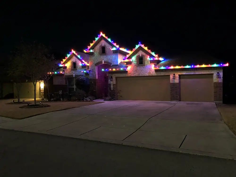 Multicolored Christmas lights outlining roofline and gables of a stone brick home at night