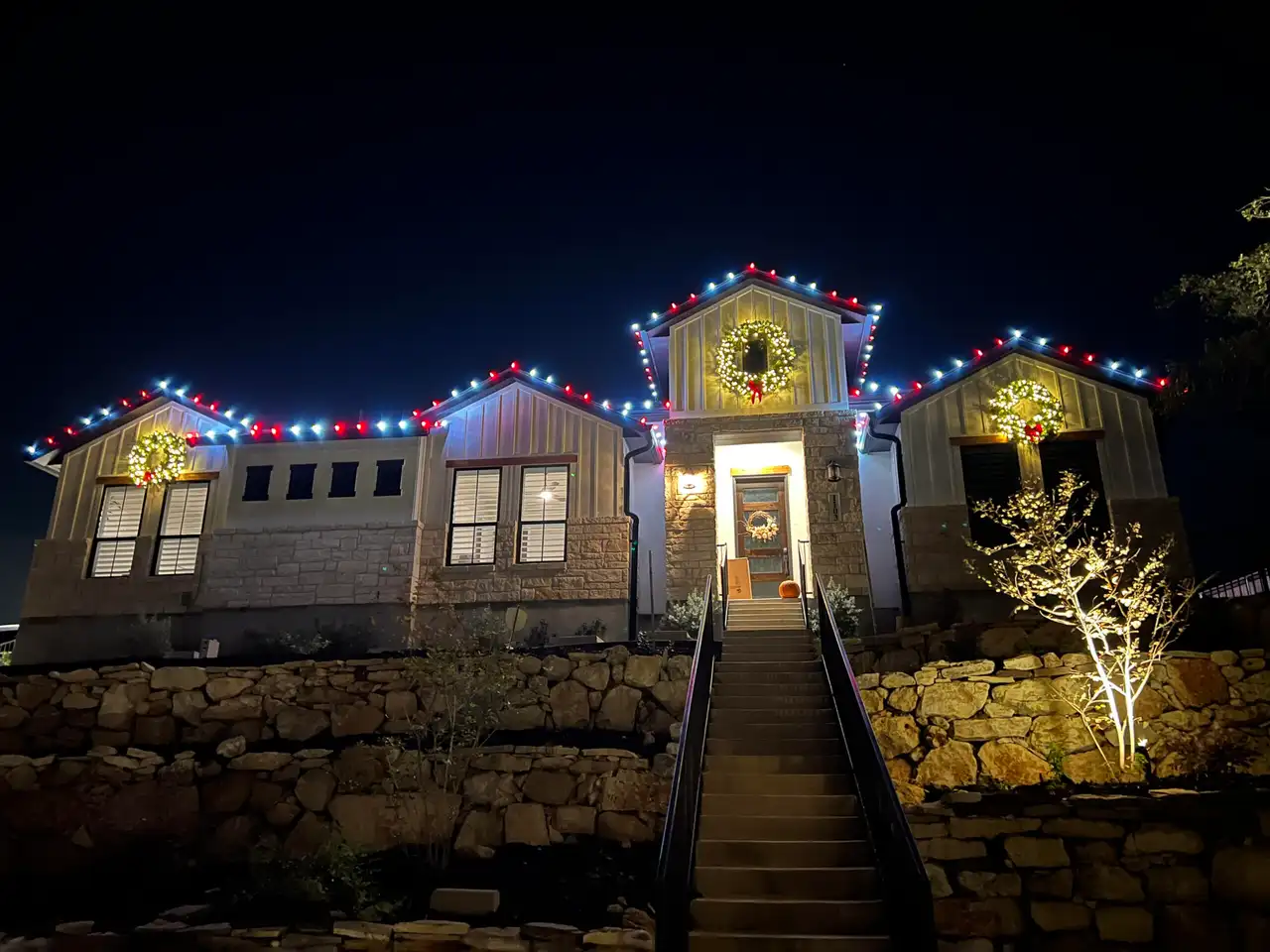 Red and white roofline Christmas lights with lit wreaths on a stone hillside home at night