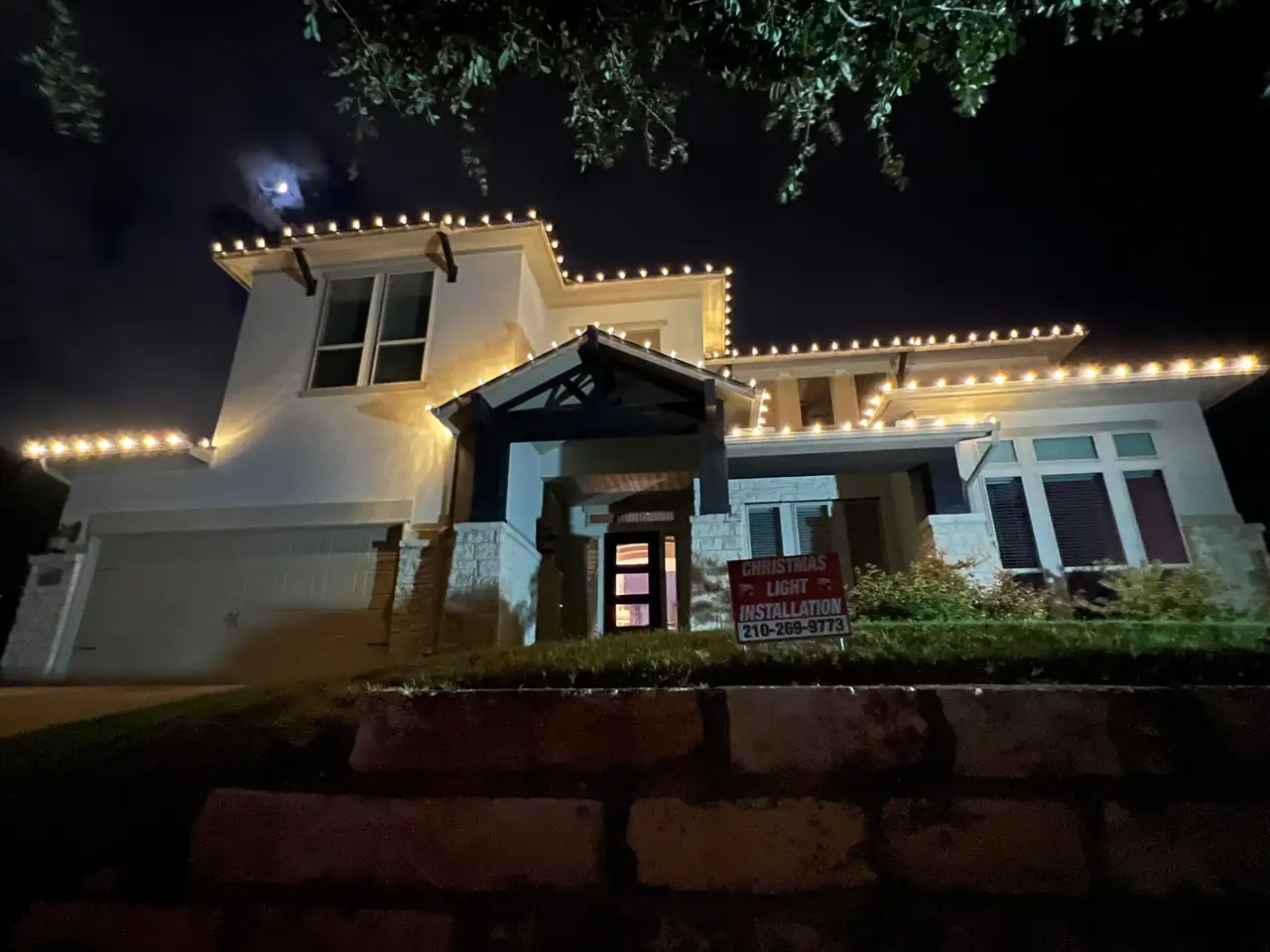 Warm white Christmas lights outlining rooflines of two-story stucco home at night