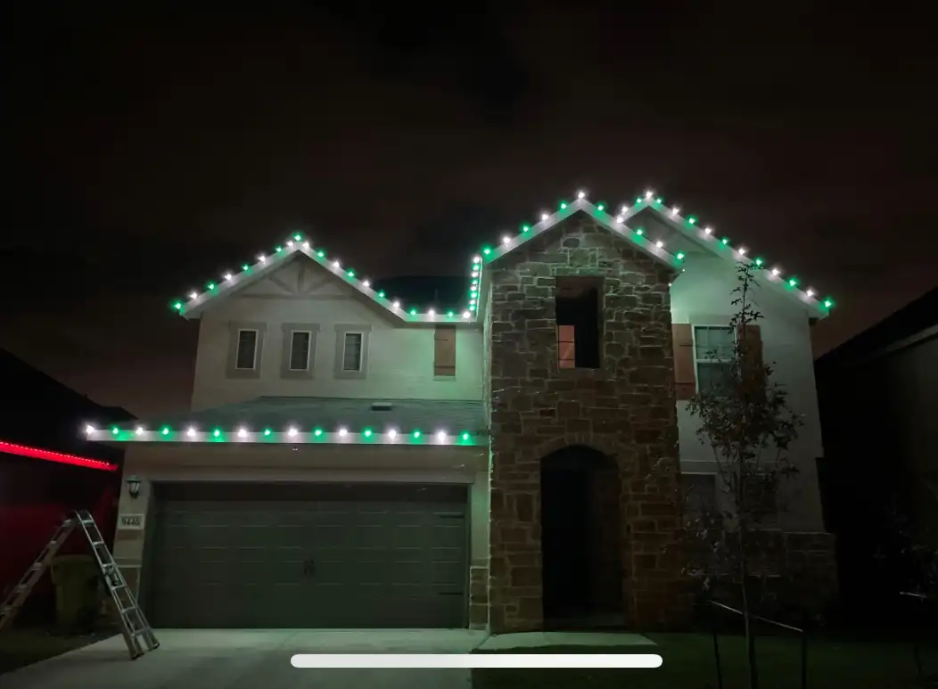 Green and white Christmas lights outlining rooflines of two-story stucco and stone home at night