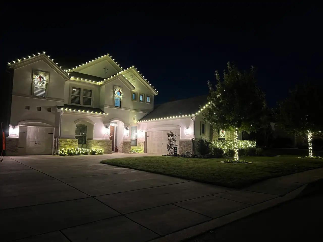Two-story stucco home outlined in warm white holiday roofline lights at night, with wreaths and wrapped tree